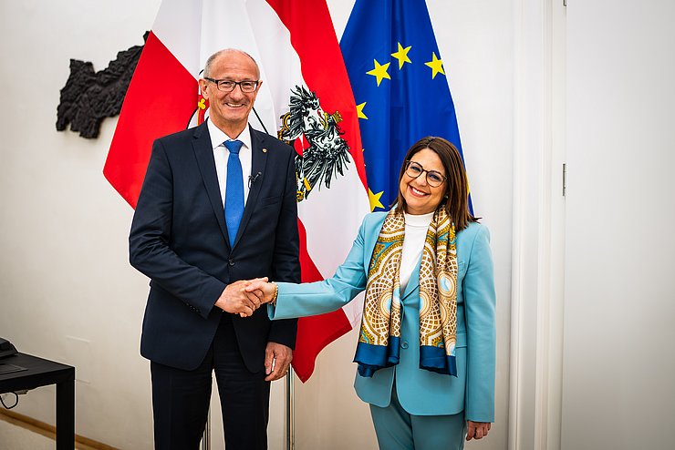 © Land Tirol Mattle und Meli Daudey handeschüttelnd vor Fahnen in Büroräumlichkeiten