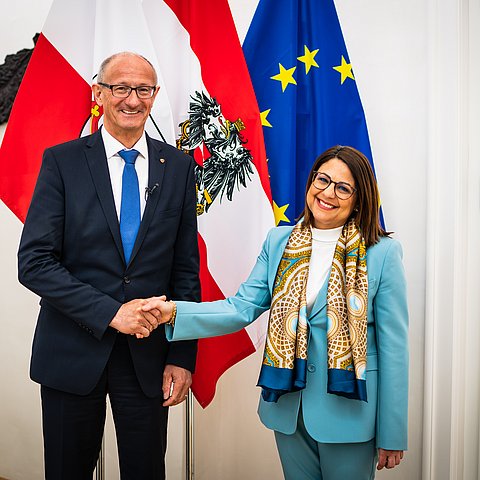 Mattle und Meli Daudey handeschüttelnd vor Fahnen in Büroräumlichkeiten