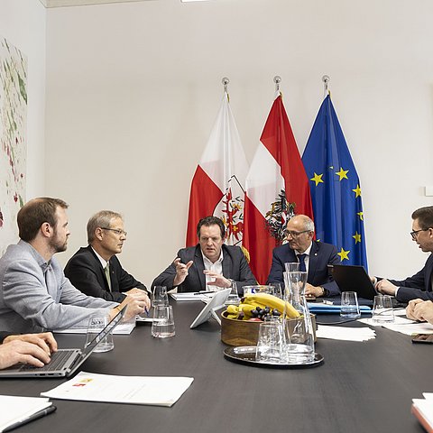 Mehrere Männer, darunter LR Zumtobel und LH Mattle, sitzen um einen Tisch herum und besprechen sich.