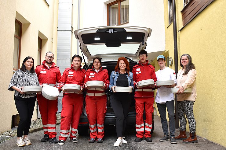 Gruppenfoto vor Auto mit geöffneten Kofferraum, Personen halten alle Essensbox in den Händen