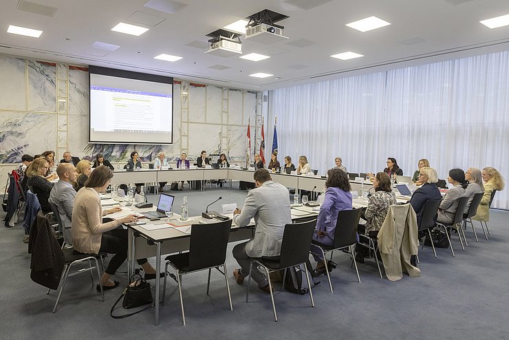 Personen sitzen um großen U-förmiger Tisch, Leinwand zeigt Folie mit Text 