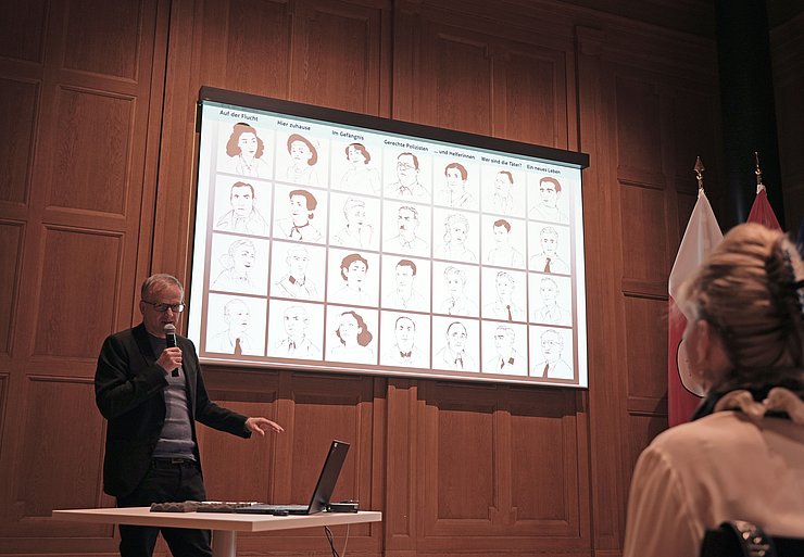 © Land Tirol/Neuner Ein Mann steht vor einer Leinwand und spricht in ein Mikro.