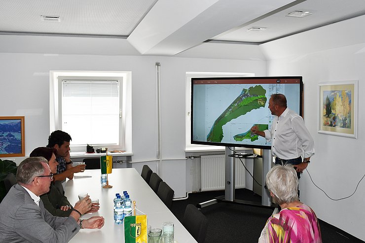 © Land Tirol/Pölzl Raum in Gemeindeamt; Bürgermeister steht vor Bildschirm mit Karte; mehrere Personen um Tisch sitzend hören ihm zu