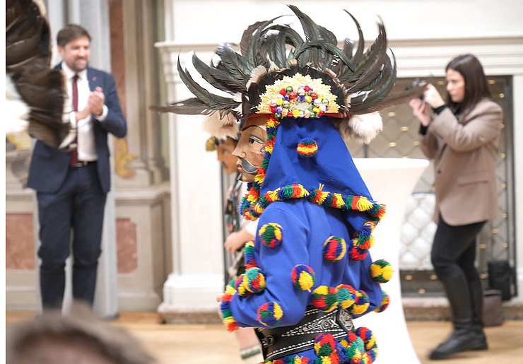 Faschingsfigur mit Holzlarve und blauem Kostüm, auf dem bunte Bälle genäht sind.