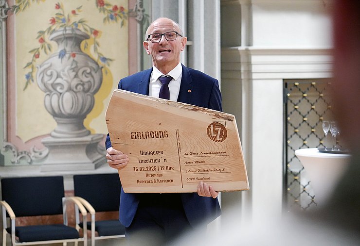 LH Mattle, wie er die Einladung, die auf einem Holzbrett geschrieben steht, in den Händen hält.