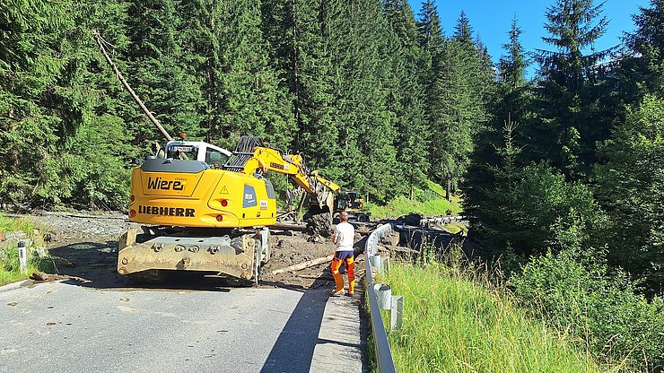 Bagger entfernt Erde/Schlamm mit Steinen und Baumstämmen von Straße.