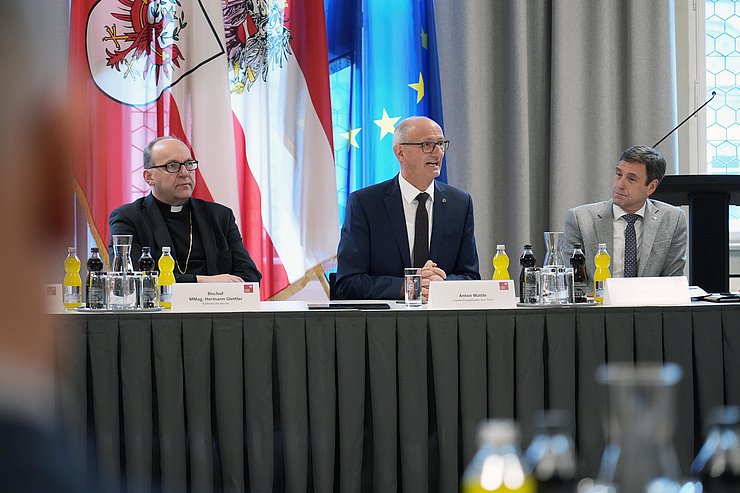 Drei Männer sitzen vor Fahnen an einem länglichen Tisch.