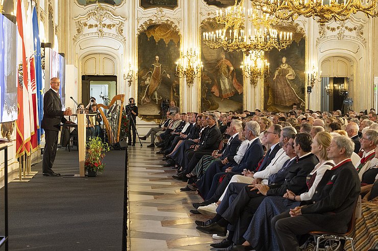 © Land Tirol/Die Fotografen Riesensaal, seitlich: LH Mattle spricht am Rednerpult, rechts die Sitzreihen mit Personen, von der Wand hängen prachtvolle Luster