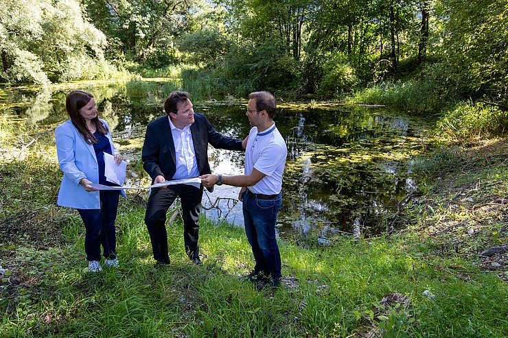 © Land Tirol Holzer, Zumtobel und Guggenberger schauen sich das Projekt auf einer Karte an. Im Hintergrund ist der Teich zu sehen.