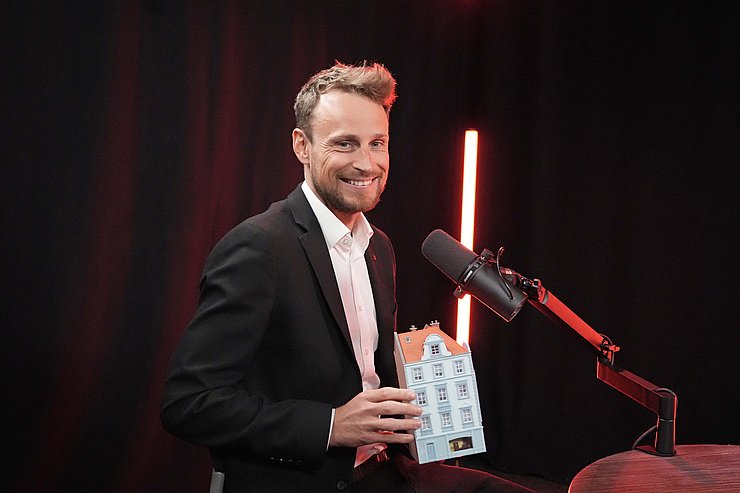 Person sitzt vor Mikrofon und hält Miniaturhaus in der Hand