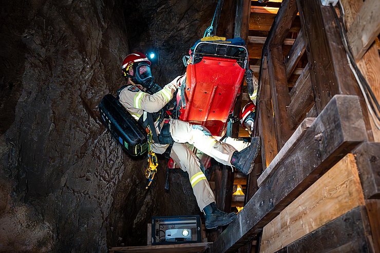 Ein Mitglied der Grubenwehr sowie die rote Trage mit verletzten werden mit einem Seil neben einer Treppe abgeseilt