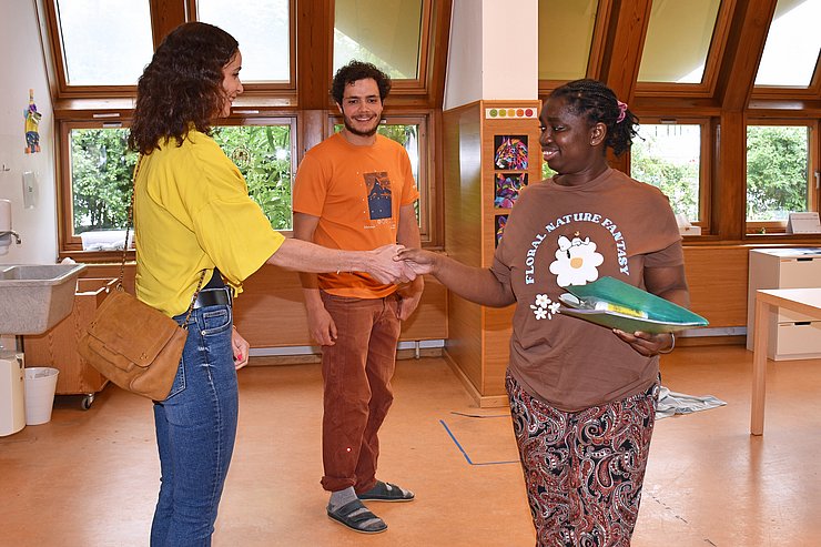 LRin reicht einer dunkelhäutigen Klientin die Hand, beide lächeln, dahinter steht Betreuer