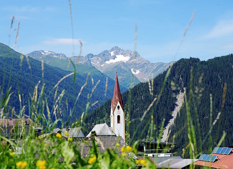 © Land Tirol/Neuner Blick durch Grashalme auf Dorf mit Kirchturm, dahinter Berg mit Schneefeldern
