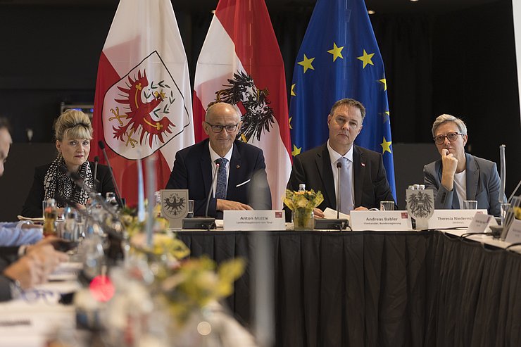 Personen an einem Tisch, im Hintergrund die Fahnen von Tirol, Österreich und der EU