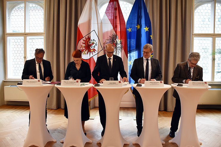 Fünf Personen stehen bei Stehtischen und unterzeichnen eine Vereinbarung