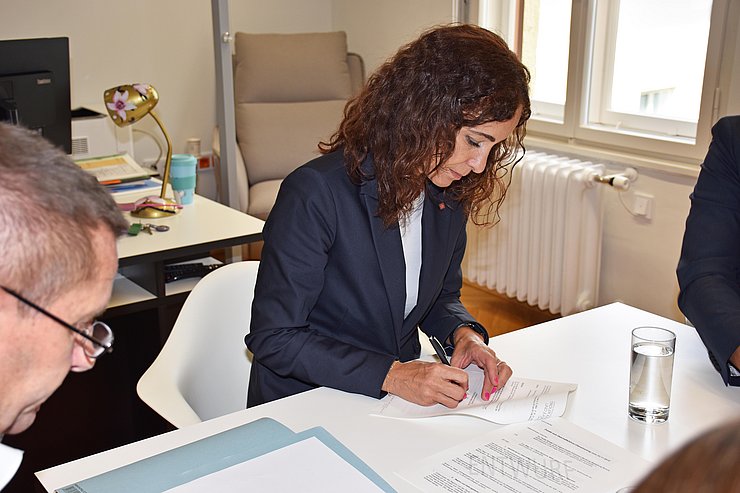 LRin unterschreibt mit Stift Formular, sitzend am Tisch, angeschnitten der Kopf des Notars