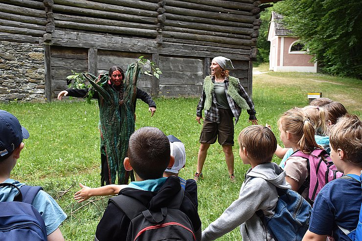 © Land Tirol/Feuersinger Zwei Märchenfiguren stehen auf einer Wiese und Kinder schauen ihnen beim Schauspielern zu.