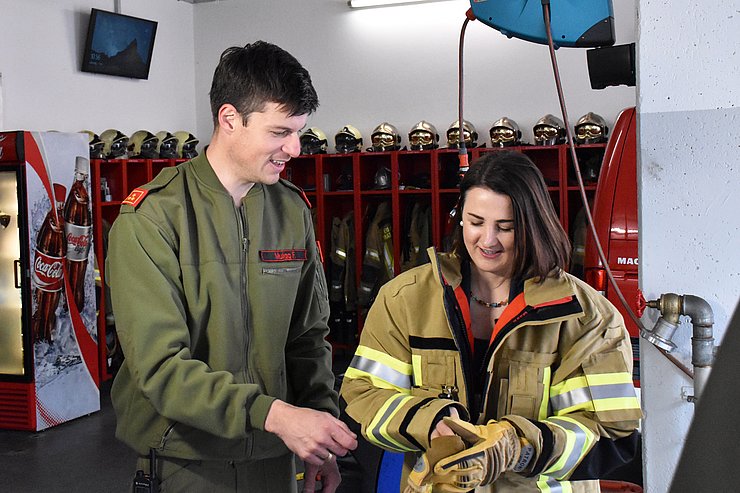 Mair rechts im Bild mit gelber Feuerwehr-Jacke mit Sicherheitsstreifen zieht sich gerade dicke Feuerwerh-Handschuhe an. Links neben ihr steht Muigg in grüner Feuerwehr-Uniform und hilft Mair beim Anziehen der Handschuhe. Beide lachen. Im Hintergrund sind rote Schränke mit der Ausrüstung der Feuerwehr zu sehen: Helme, jacken, Schuhe und Handschuhe