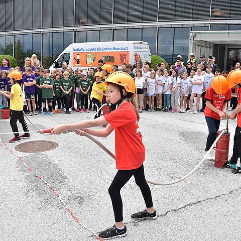 Foto Lösch-Übung: Ein Kind in einem roten T-Shirt hat einen Feuerwehrschlauch in der Hand und spritzt Wasser auf ein nicht erkennbares Ziel. Dahinter stehen zwei weitere in roten T-Shirts und pumpen Wasser in den Schlauch. 