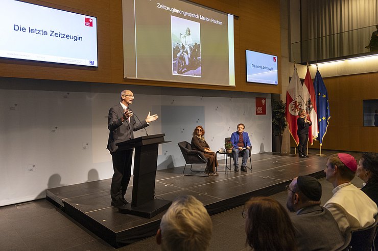 © Land Tirol/Die Fotografen Am 9. November 2023 begrüßte LH Anton Mattle (stehend links) die Zeitzeugin Marion Fischer (sitzend Mitte) im Großen Saal im Landhaus, die vor Publikum ein Gespräch mit dem Zeithistoriker Horst Schreiber (sitzend rechts) von ERINNERN:AT führte. Ganz rechts steht auf dem Podium eine Gebärdendolmetsch.