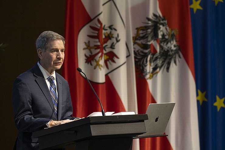 © Land Tirol/Die Fotografen Ein Mann steht vor Fahnen an einem Rednerpult.