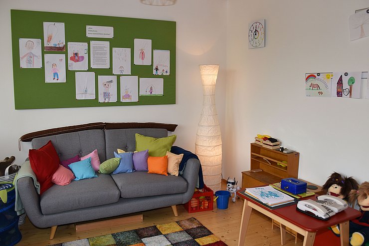 Ein Spielzimmer mit Sofa, bunten Kissen und einer Pinnwand mit Kinderzeichnungen. 