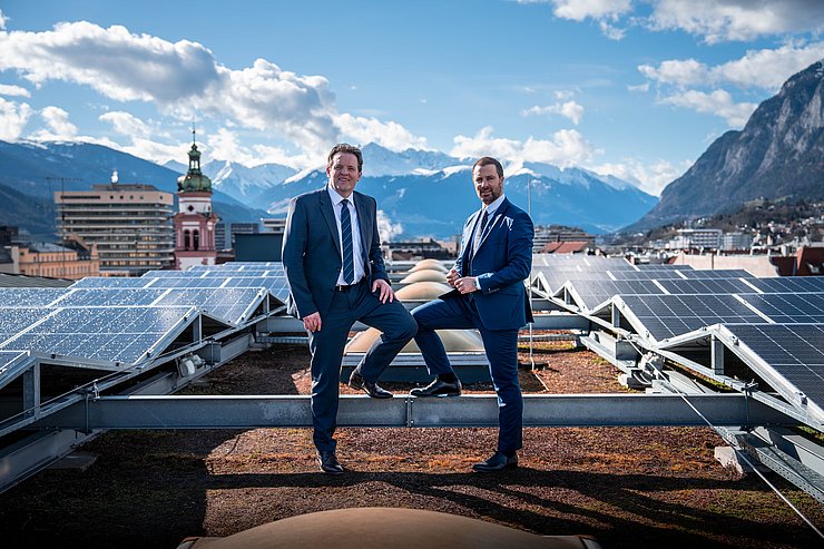 Zumtobel und Dornauer auf Dach Landhaus; im Hintergrund Innsbruck; am Dach PV-Anlagen; beide stehen mit jeweils einem Fuß auf Stahlträger der Anlage