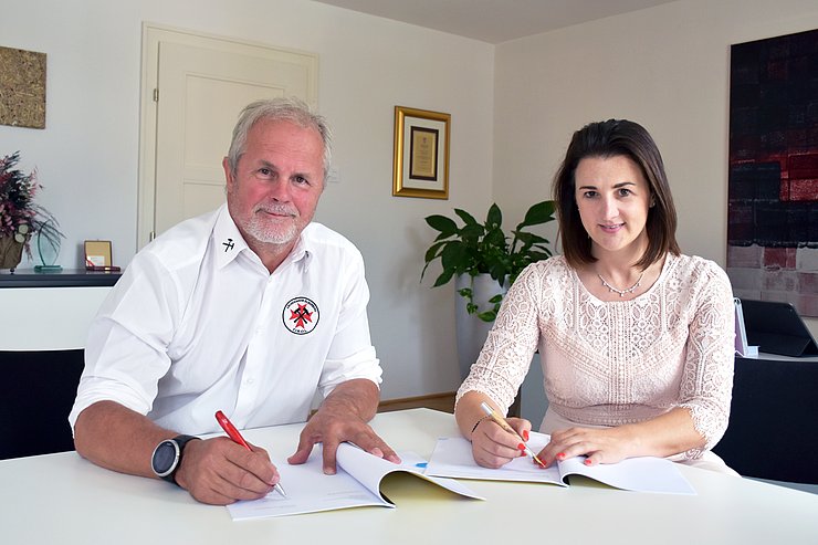 Höfer und Mair sitzen in einem Büro an einem Tisch; vor ohnen zwei Mappen; beide halten Stifte in den Händen