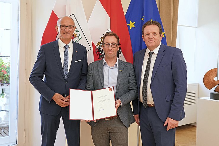 Drei Menschen posieren vor mehreren Flaggen.