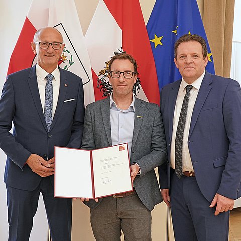 Drei Menschen posieren vor mehreren Flaggen.