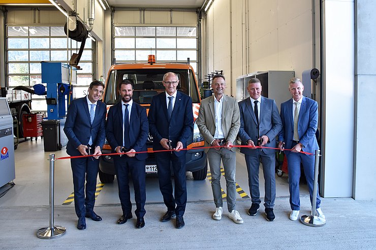 Mehrere Männer in Anzügen hinter einem roten Band, welches sie durchschneiden; im Hintergrund eine Werkstatt