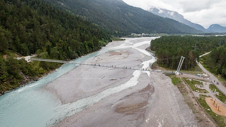 Auf dem Bild ist eine große Hängebrücke über dem frei fließendem Lech der in dem Bild einen niedrigen Wasserstand aufweist.