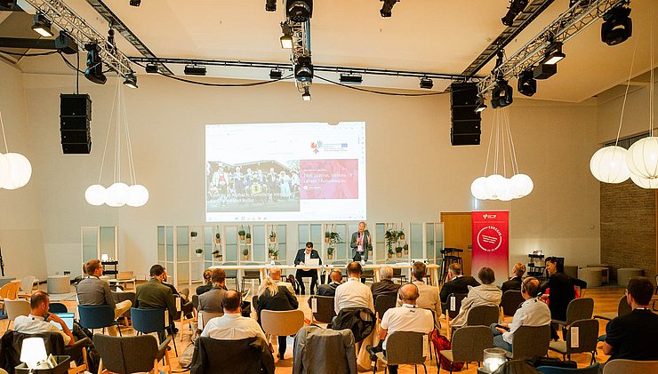 Saal mit Stuhlreihen auf denen Menschen sitzen