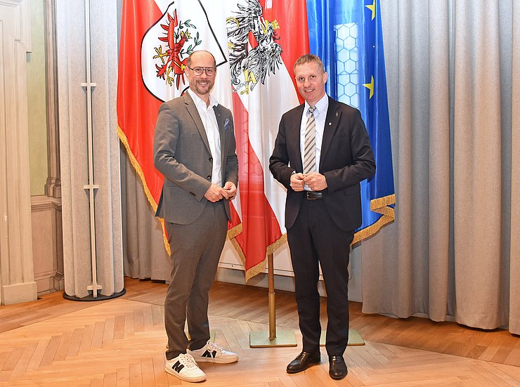 © Land Tirol/Pichler LR Gerber und LR Walcher zusammen vor einer Österreich-, einer Tirol- und einer EU-Flagge.