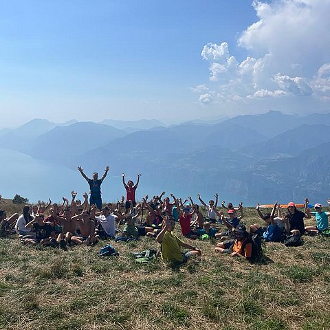 Gruppe Jugendlicher streckt Hände nach oben, sitzt auf Wiese - dahinter Bergkulisse und See