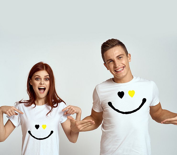 © Land Tirol Eine Frau und ein Mann lachen in die Kamera und zeigen auf ihr weißes T-Shirt mit einem Smiley darauf.