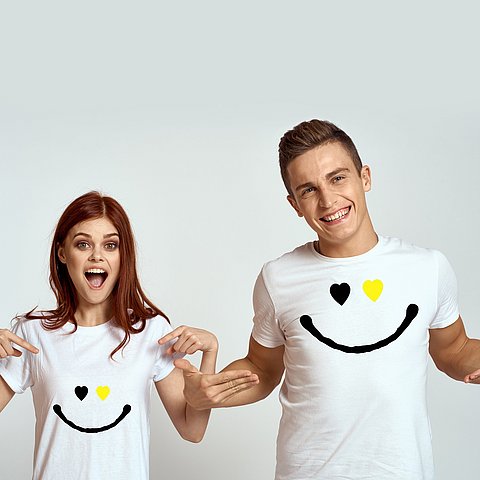 Eine Frau und ein Mann lachen in die Kamera und zeigen auf ihr weißes T-Shirt mit einem Smiley darauf.