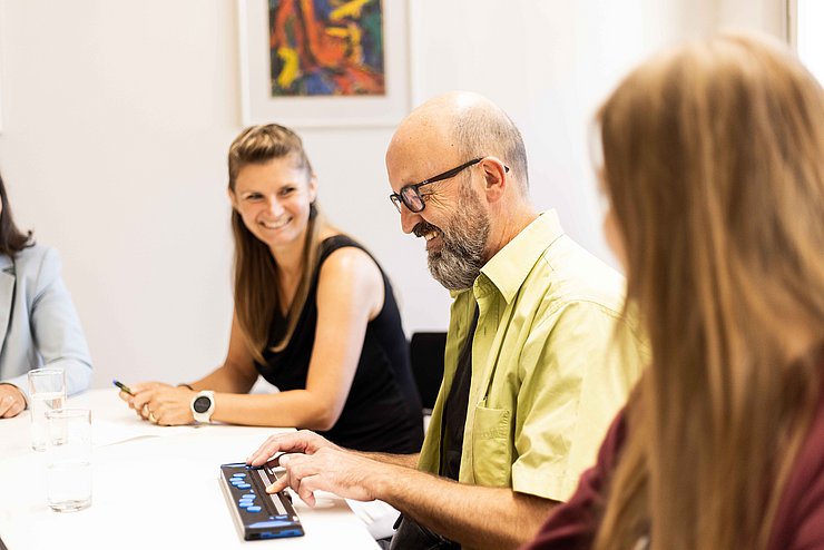 drei Personen am Tisch, eine Person verwendet Braillezeile