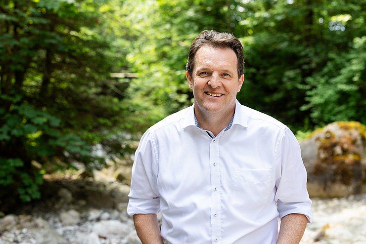 Ein Mann steht in weißem Hemd und mit hochgekrempelten Ärmeln vor Bäumen.