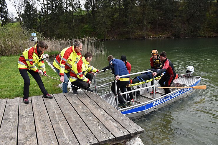 © Land Tirol/Pölzl Das Boot der Wasserrettung ist am Steg angekommen: 4 Männer mit Neoprenanzügen stehen im Boot und halten eine Trage. Auf dieser liegt die Verunglückte. Am Steg stehen drei Mitglieder der Wasserettung mit Gelb-Roten Jacken. Sie halten das Boot fest und nehmen die Verunglückte auf der Trage entgegen.