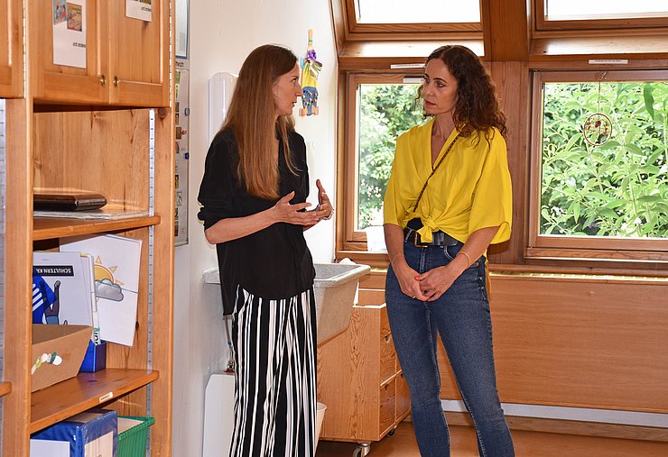 LRin unterhält sich mit GFin. diese gestikuliert, sie befinden sich in Raum mit Holzregal und Fenstern