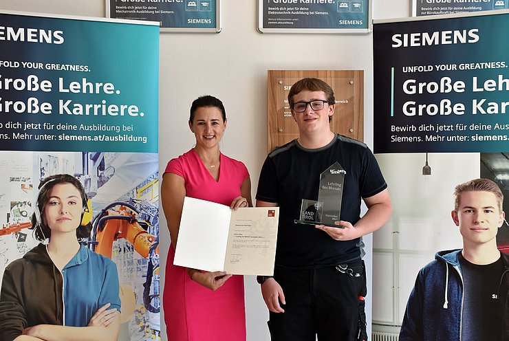 Mair und Huber stehen nebeneinander; Mair hält eine Ukrunde, Huber eine Auszeichnung aus Glas mit Aufschrift Lehrling des Monats