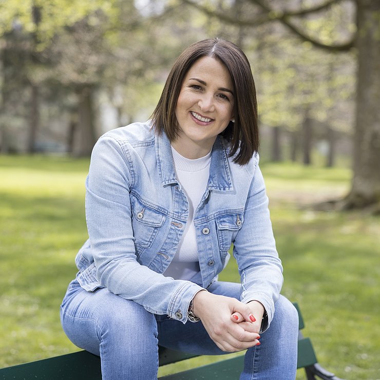 © Land Tirol/Sedlak Mit einem Jeansanzug bekleidet sitzt Jugendlandesrätin Mair sitzt auf der Rückenlehne einer dunkelgrünen Parkbank, die von Bäumen umgeben ist.