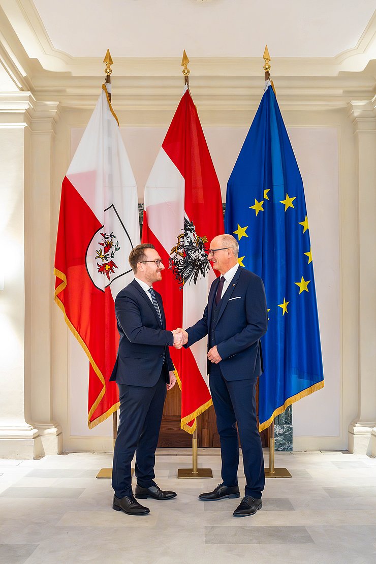 Zwei Männer schütteln sich vor Fahnen die Hand.