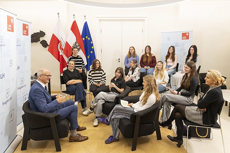 © Land Tirol/Die Fotografen Einige Schülerinnen setzen auf Stühlen und Blicken auf den Landeshauptmann, der vor Roll-ups sitzt.