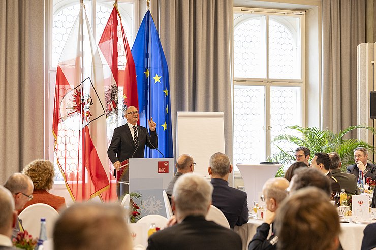 Mattle sprechend; Personen an runden Tischen sitzend und zuhörend