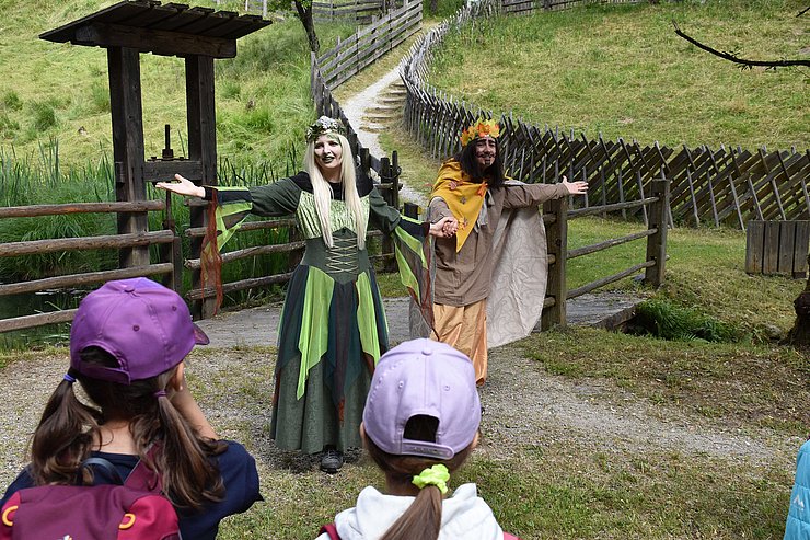 © Land Tirol/Feuersinger Zwei Märchenfiguren stehen auf einer Weggabelung und Kinder schauen ihnen beim Vorführen zu.