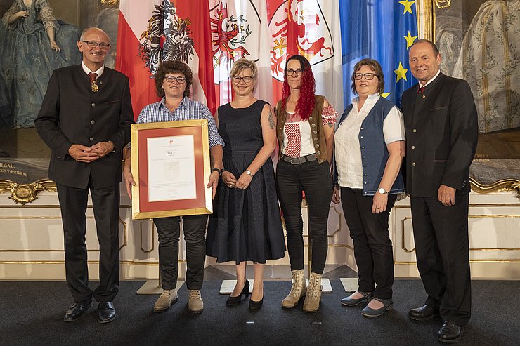 © Land Tirol/Die Fotografen links LH Mattle, in der Mitte die Famile bestehend aus vier Frauen, rechts LR Geisler, eine Frau hält golden gerahmte Urkunde in den Händen