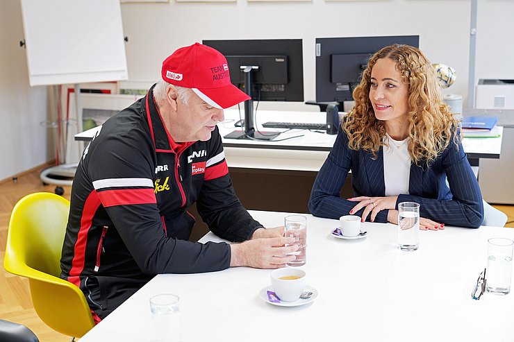 Eine Frau spricht an einem Tisch sitzen mit einem Mann in Trainingsanzug.
