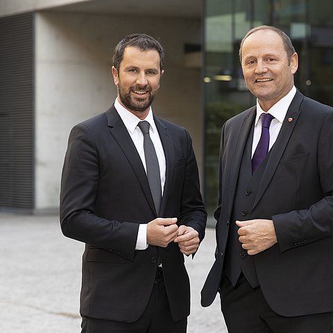 Dornauer und Geisler stehen in dunklen Anzügen nebeneinander und lächen in die Kamera